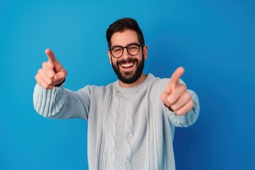 Young handsome man with beard wearing casual sweater and glasses over blue background, pointing to the camera with fingers, smiling positive and cheerful.