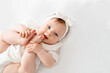 © Any Grant - Newborn baby girl plays with her legs pushing them into her mouth in white clothes on the bed at home on her back, top view, space for text