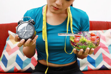 Asian women is on dieting time looking at clock. Holding vegetable salad bowl.