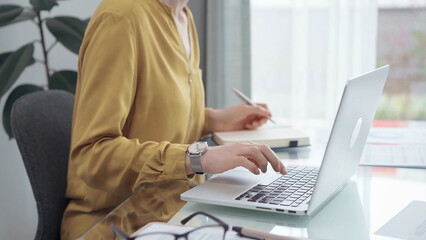 Unrecognizable professional woman is making notes and working with laptop in modern office. Yellow blouse is passing to adult businesswoman. Business concept