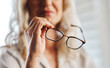 © HockleyM3/peopleimages.com - Business, woman and hands with glasses in home for vision, bluelight and lens for eye care. Zoom, freelancer and closeup of entrepreneur with eyewear for clear sight, frame design and help to see