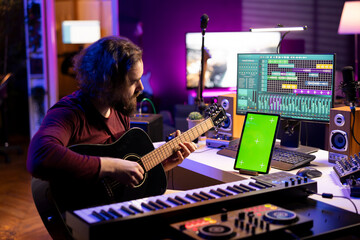 Leinwandbilder - Sound engineer artist playing acoustic guitar in his home studio, recording new music with daw software on computer. Musician learns new instrument accords next to greenscreen on tablet.
