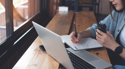 Canvas Print - Business woman sitting on wooden table in coffee shop, drinking coffee, writing on notebook, working on laptop computer, close up. Female student studying online, e-learning, work from cafe