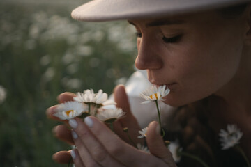 Wall Mural - A woman is smelling a bunch of white flowers. The flowers are in a field and the woman is wearing a hat.