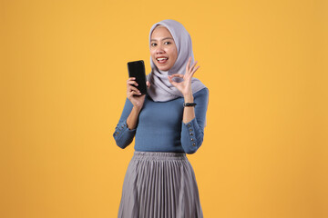 happy asian indonesian muslim woman holding smart phone and showing smart phone blank screen and giving ok finger gesture on isolated yellow background