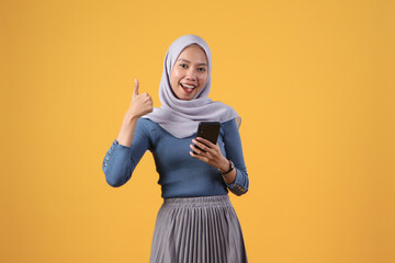 Wall Mural - asian indonesian muslim woman holding smart phone giving thumb up finger gesture on isolated yellow background