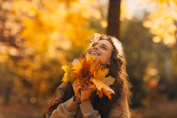 Wall Mural - Smiling young woman enjoying the autumn season in the fall park with the yellow leaves at sunset