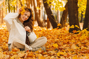 Wall Mural - Smiling young woman sits in the autumn foliage on the ground of the forest with the yellow leaves