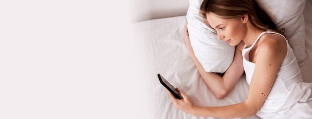 young woman wearing white pajama lying in bed use mobile phone rest relax spend time in her bedroom, top view