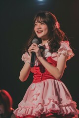 Wall Mural - A photo of an extremely beautiful young Japanese girl, wearing pink and red short dress with frills on the shoulders, holding microphone in hand, dancing at school stage, white skin tone, dark brown h