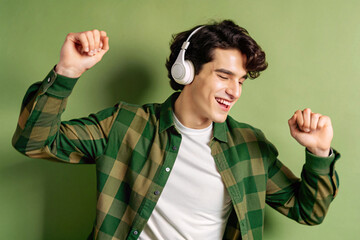 A cheerful man with dark curly hair, wearing a green plaid shirt and white headphones, dances energetically to music against a green background