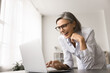 © fizkes - Happy mature 50s businesswoman with glasses leaned at desk using laptop, look at device screen and laughing, having positive and enjoyable on-line interaction, get great news, sales growth, promotion