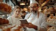 © The Little Hut - Caucasian handsome man baker in uniform showing something on tablet device to woman colleague Bakers talking and discussing work with computer in hands Bakery concept Workday at bakesh : Generative AI