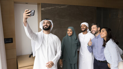 Wall Mural - Happy young Arabian project leader man taking selfie with Middle Eastern and European business partners after meeting, holding smartphone, standing in office with diverse team behind