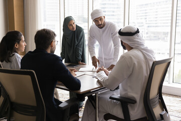 Wall Mural - Business team of young serious Arabic managers discussing teamwork, project strategy with Arabian boss and European partners at meeting, standing at table, speaking to sitting colleagues