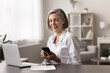 © fizkes - Pretty middle-aged businesswoman using new mobile application in office. Woman sit at desk with laptop, holds modern gadget, check messages, read good news in e-mail, get positive feedback from client