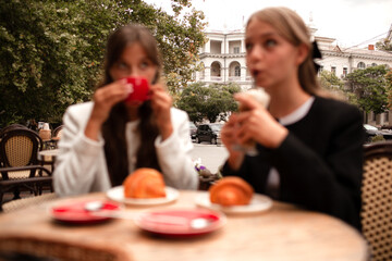 Two beautiful young girls are sitting in a French cafe, eating a croissant and drinking coffee with whipped cream. Teenagers are dressed in business style, black and white jacket in Old Money style