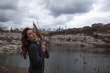 A beautiful woman dressed in gray pants and a sweater walks against the background of a career and lake in stylish clothes