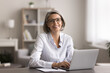 © fizkes - Happy middle-aged woman with short gray hair and glasses, smile, look at camera, seated in cozy home office setting, working on laptop. Freelance and telework using modern tech and internet connection