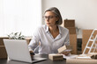 © fizkes - Woman focused on her laptop while hold small package in modern office setting filled with cardboard boxes, engaged in logistical, shipping task, overseeing inventory, packaging, dispatch operations