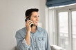 © fizkes - Cheerful handsome young Hispanic man in casual talking on cellphone indoors, enjoying telephone call communication, looking away, speaking on mobile phone at home, smiling, laughing