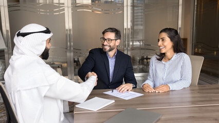 Wall Mural - Cheerful European married couple shaking hands with Arabian business consultant, sitting at office meeting table, smiling, discussing property investment, real estate buying. Banner shot