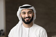 © fizkes - Positive handsome Arabic businessman with beard wearing keffiyeh and thobe, looking at camera, smiling. Successful company owner, young Arab business leader man head shot video call screen portrait