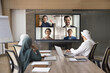 © fizkes - Group of Arab business owners meeting at large table in boardroom, talking on group video call, conference chat, looking at split screen with team of foreign colleagues, partners