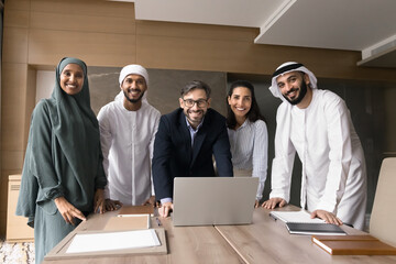 Wall Mural - Diverse Caucasian and Middle Eastern business team posing at laptop, looking at camera, smiling, standing at meeting table. Young Arabian and European group with middle aged Hispanic leader portrait