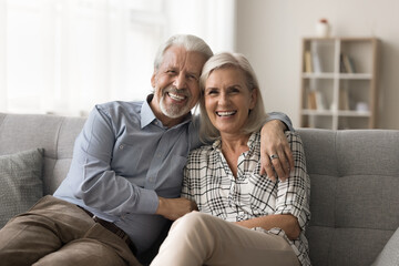 Portrait of attractive aged retired couple hugging resting together at home sit on sofa laughing looking at camera, enjoy their harmonic relationships lifelong marriage and carefree life on retirement