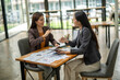 © bunyiam - Two women sitting at a table talking and looking at papers