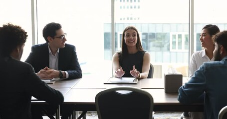 Wall Mural - Positive confident young business leader woman talking to diverse team of colleagues, brainstorming at meeting table in office, discussing project, teamwork strategy with partners, speaking, smiling