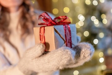 Gift, Winter, Christmas - Woman holding a wrapped present with red ribbon in her gloved hands, standing in front of a Christmas tree with lights.