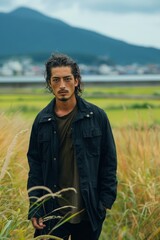 Portrait of man standing in field with mountains in background. He wears black jacket and looks directly into camera, resilience and determination. Concept of solitude and connection with nature