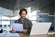 © insta_photos - Happy busy young business man accountant reading file report documents checking data online looking at laptop computer paying financial loan analyzing invoice or banking statement working in office.