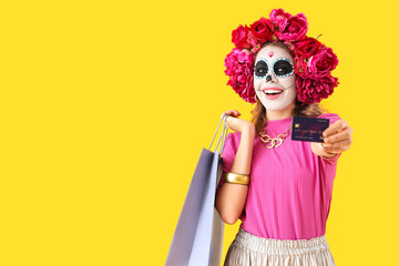 Canvas Print - Young woman with credit card and shopping bag on yellow background. Mexico's Day of the Dead (El Dia de Muertos) celebration