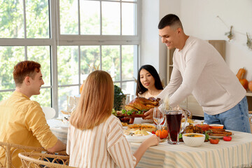 Canvas Print - Young man bringing tasty turkey to dining table with his friends at home on Thanksgiving Day