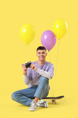 Poster - Portrait of young man with colorful balloons, photo camera and skateboard on yellow background