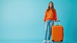 © munawaroh - A young woman in an orange sweater stands beside an orange suitcase against a blue background.