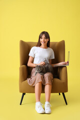 Poster - Beautiful young happy woman with cute cat and laptop sitting on armchair against yellow background