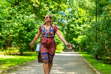 Wall Mural - Beautiful blonde woman wearing colorful floral flowing dress walking with hands spread out on green background in city park on sunny day. Front view