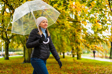 Wall Mural - Middle aged woman wearing black jacket, jeans and cap walking with transparent umbrella in city park on autumn rainy cool day. Side view