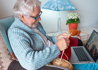 Canvas Print - Senior man at home follows online knit tutorial to relax enjoying resting home leisure activity sitting on armchair - male people knitting with computer class help - people inner lifestyle hobby