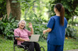 © kamonrat - Elderly woman in a wheelchair waving with a smile while holding a laptop, engaging warmly with a nurse in a green park.