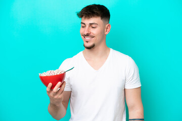 Young caucasian man holding a bowl of cereals isolated on blue background with happy expression