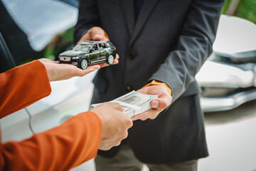 Two Asian individuals discuss a car insurance claim with an agent in front of a damaged vehicle. They review documents, discuss the policy, and address the crash and repair costs.