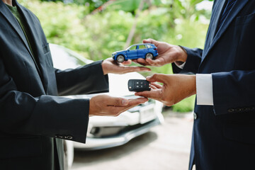 Two Asian individuals discuss a car insurance claim with an agent in front of a damaged vehicle. They review documents, discuss the policy, and address the crash and repair costs.