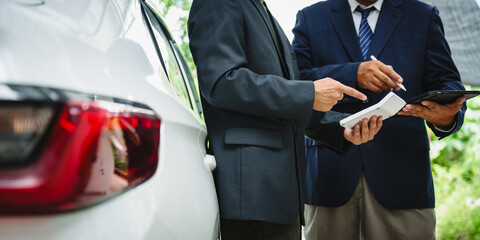 Two Asian individuals discuss a car insurance claim with an agent in front of a damaged vehicle. They review documents, discuss the policy, and address the crash and repair costs.