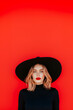 © Erik González - A woman wearing a black hat and red lipstick stands in front of a red background