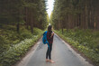 © Fauren - Back view of a woman with a blue backpack walking barefoot down a peaceful forest road, surrounded by lush greenery and tall trees, symbolizing adventure and tranquility in nature